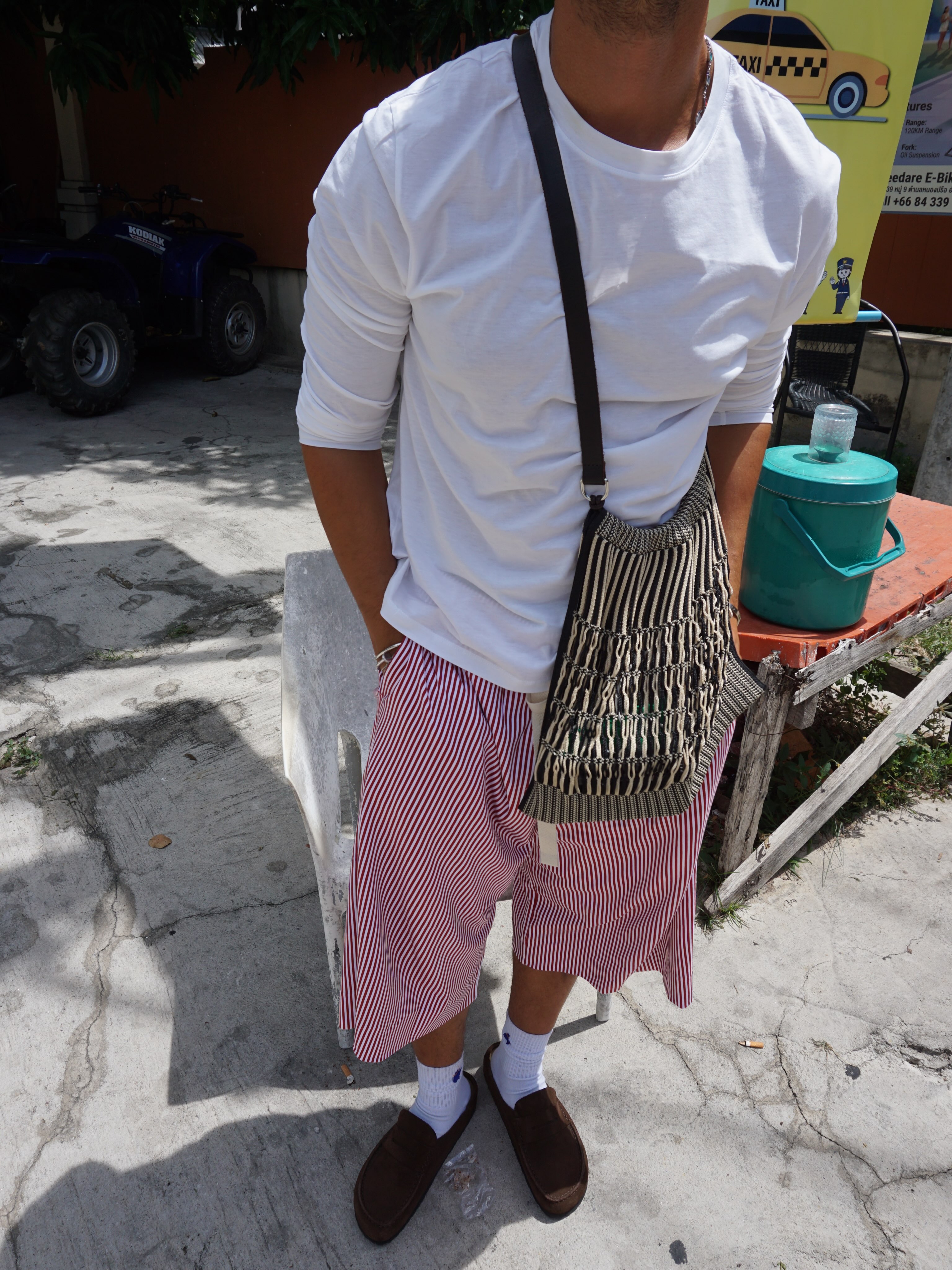 Man outdoors in a white long-sleeve top, patterned red trousers, and a crossbody bag.