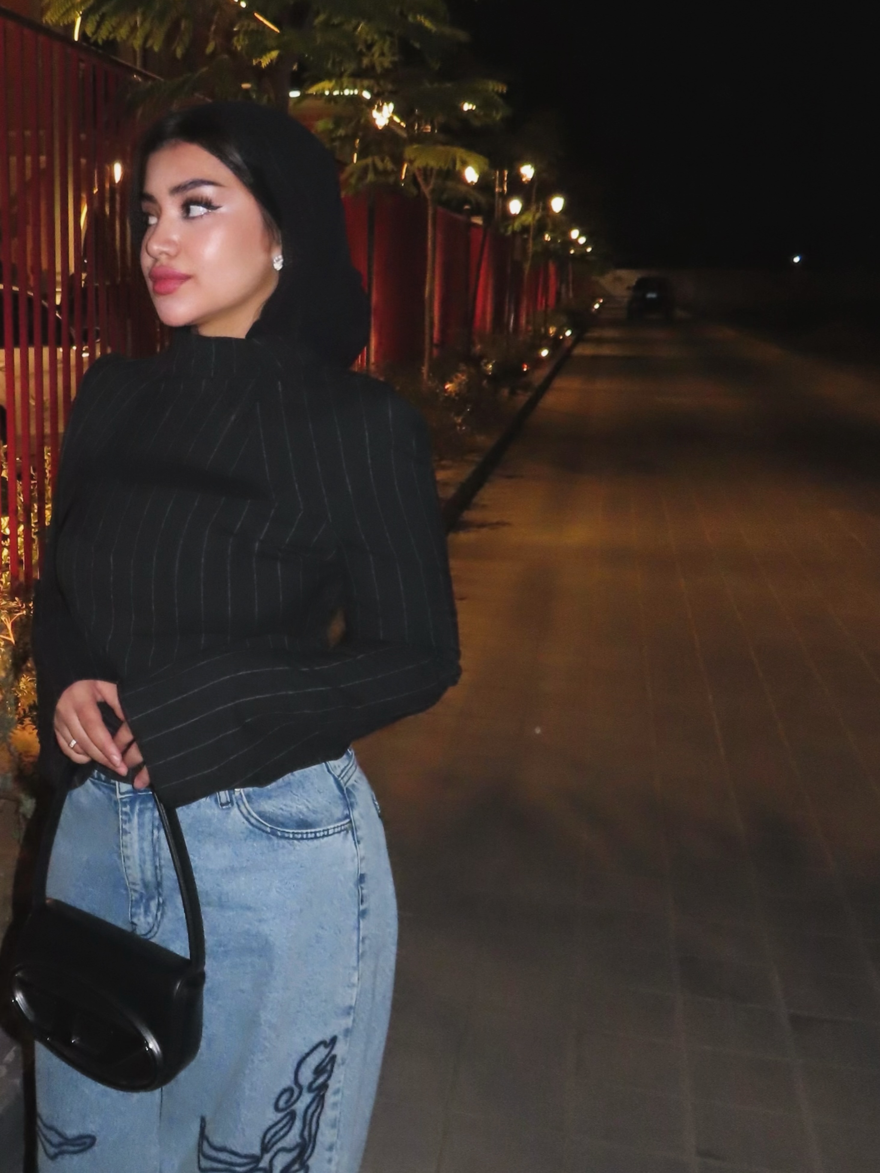 Woman in a black sweater and light blue jeans holding a black shoulder bag at night.